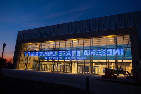 An exterior photo of Indiana State University’s Hulman Center arena in the evening. The building has clear glass windows in the front with “Indiana State University Hulman Center” in blue lettering, illuminated. A light post is visible to the right against the darkening sky.